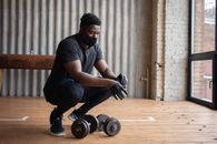 Black sportsman in mask hunkering down near dumbbells in gym