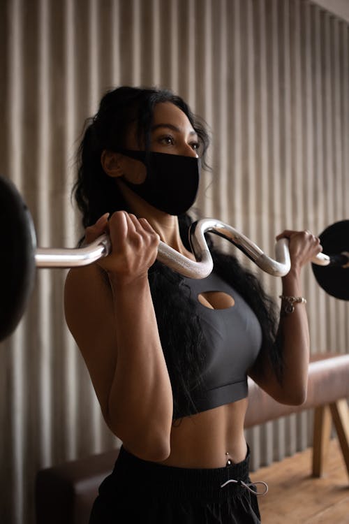 Young sporty female in activewear and face mask exercising with barbell in gym
