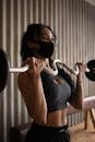 Young sporty female in activewear and face mask exercising with barbell in gym