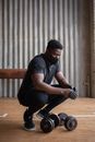 Full length focused African American sportsman in activewear and mask putting on latex gloves while hunkering down near dumbbells in gym