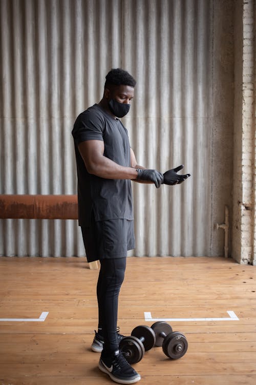 Side view of African American sportsman in protective mask and gloves standing near dumbbells while training in gym
