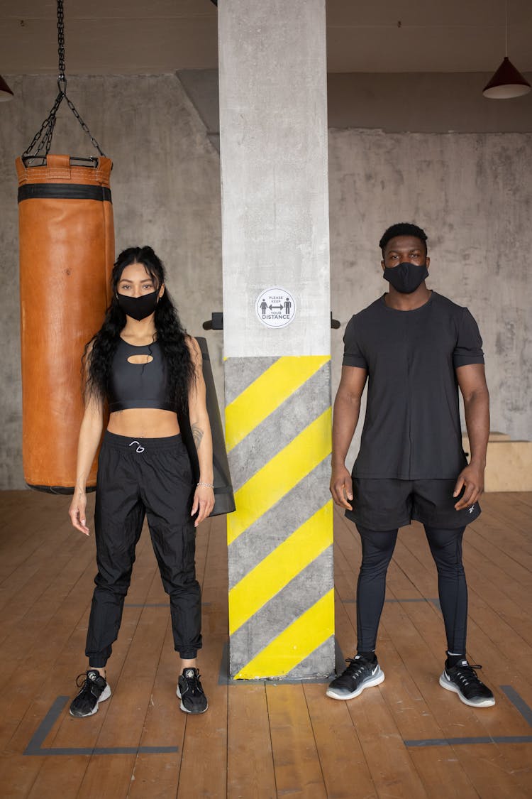 Confident Multiracial Sportive Couple In Masks Standing Near Boxing Bag