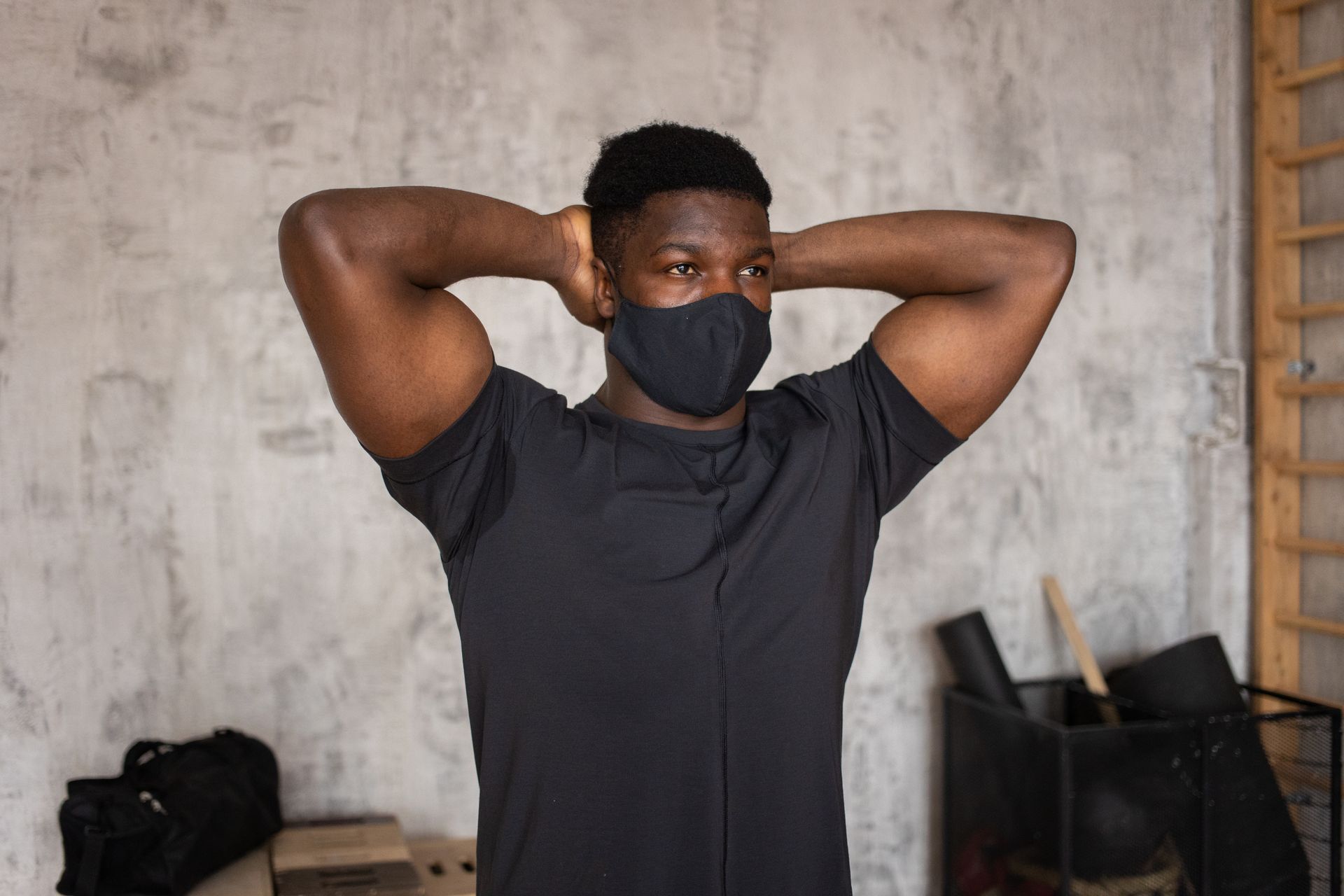 Serious African American male with short dark hair in protective sterile mask standing with raised arms and looking away against concrete wall