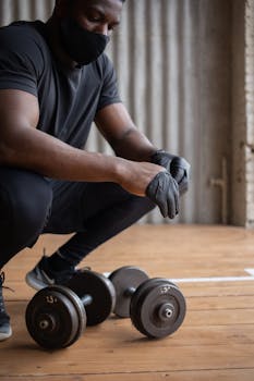 Crop black sportsman in mask squatting down and wearing gloves while training with dumbbell in gym in daytime