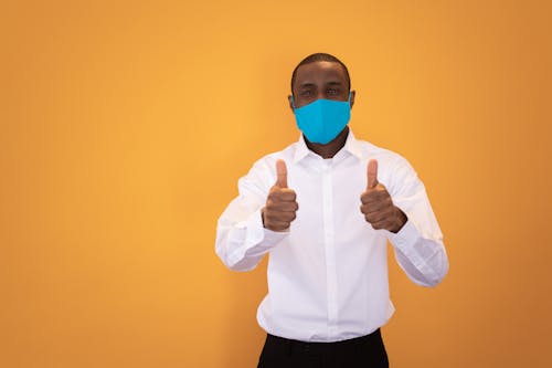 Positive African American male in protective mask and shirt standing and showing thumbs up gesture against yellow background in studio