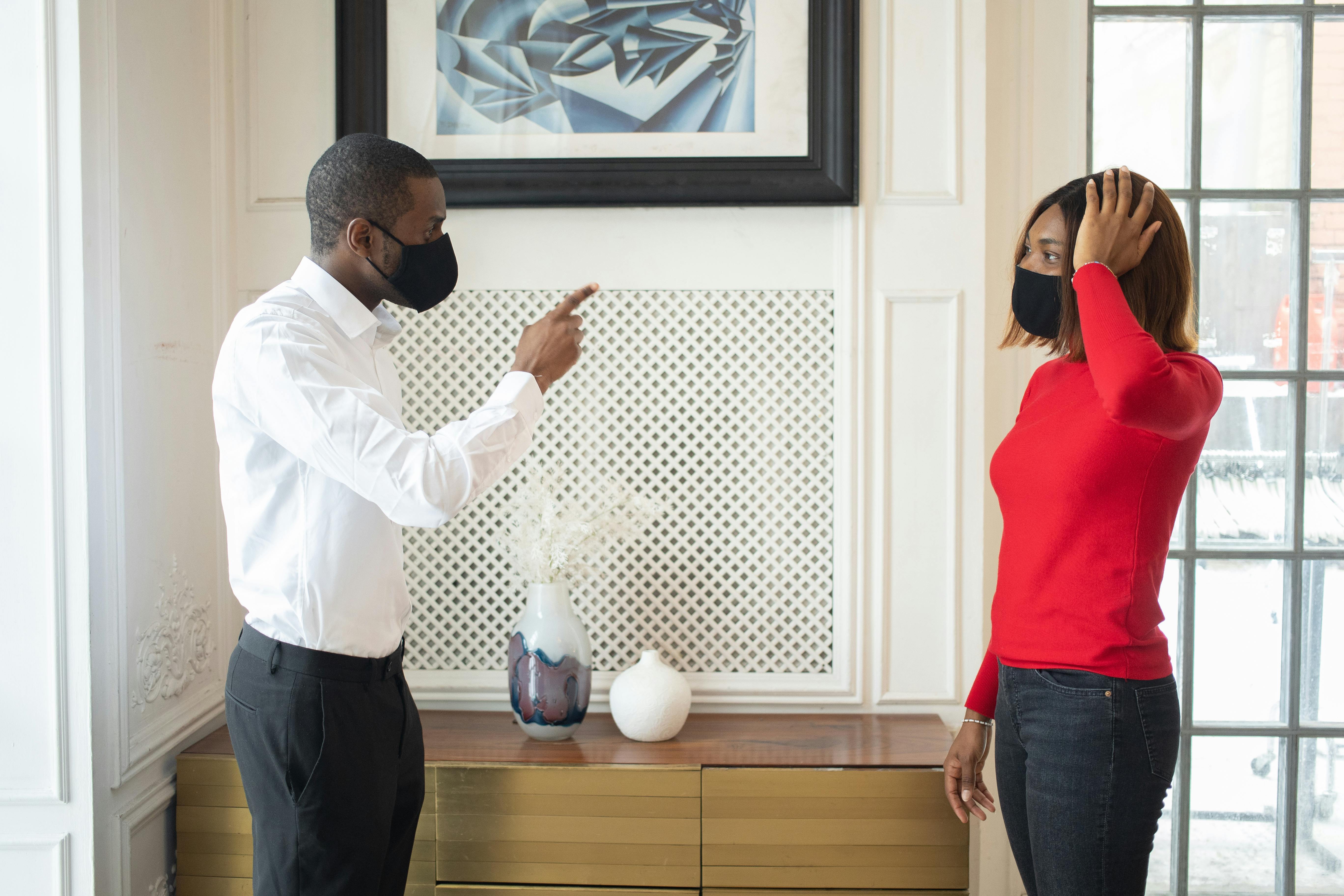 Side view of African American couple in protective masks and casual clothes standing and quarreling in light room in daytime