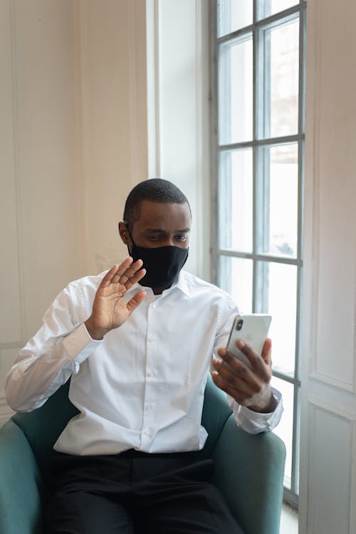 Calm African American male in formal clothes sitting on armchair near window in light room and talking on video call on smartphone in daytime