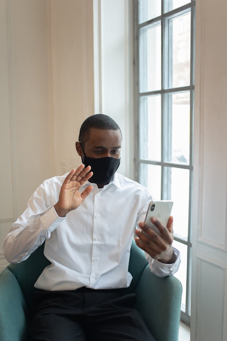 Black Man In Mask Talking On Video Call On Smartphone In Light Room