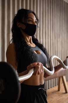A determined woman lifting a curl bar indoors, wearing sportswear and a protective mask.