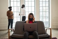 Ethnic female worker in fabric mask with netbook and thumb up speaking on cellphone against male partners with takeaway coffee