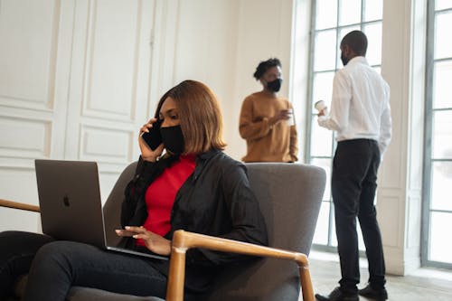 Free Black female worker in fabric mask with netbook speaking on mobile phone against unrecognizable male colleagues with disposable coffee in workspace Stock Photo