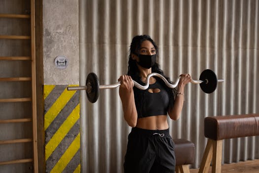 Fit African American female in fabric mask and sportswear looking forward during training with curl bar in gym