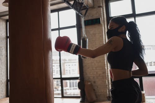 Tattooed black boxer in face mask punching heavy bag indoors