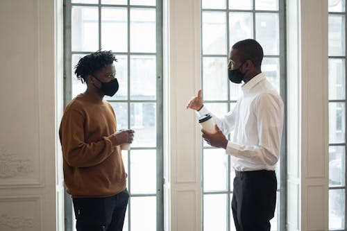 Side view of African American male executive in fabric mask with disposable hot beverage speaking with coworker while looking at each other indoors