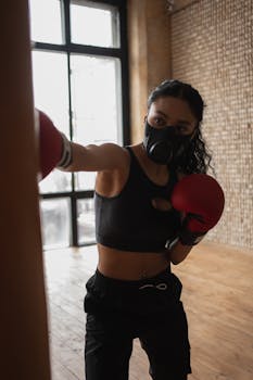 Concentrated African American female fighter in sportswear and fabric mask punching boxing bag during training in gymnasium
