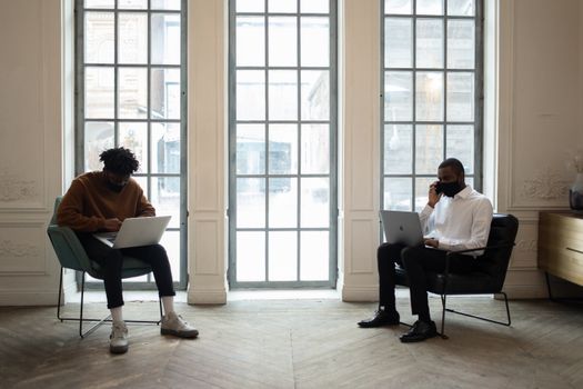 Ethnic male executive in cloth face mask talking on cellphone against colleague with netbook in armchair against windows in workspace