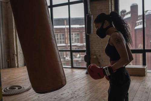 Side view of ethnic female boxer in textile mask against heavy bag during workout in gym