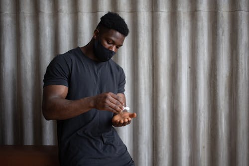 Free Black male athlete in sports clothes and textile mask applying antiseptic gel on hand against ribbed gray wall Stock Photo