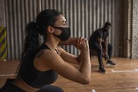 Tattooed black sportswoman in textile mask squatting against male partner with dumbbell during workout in gymnasium