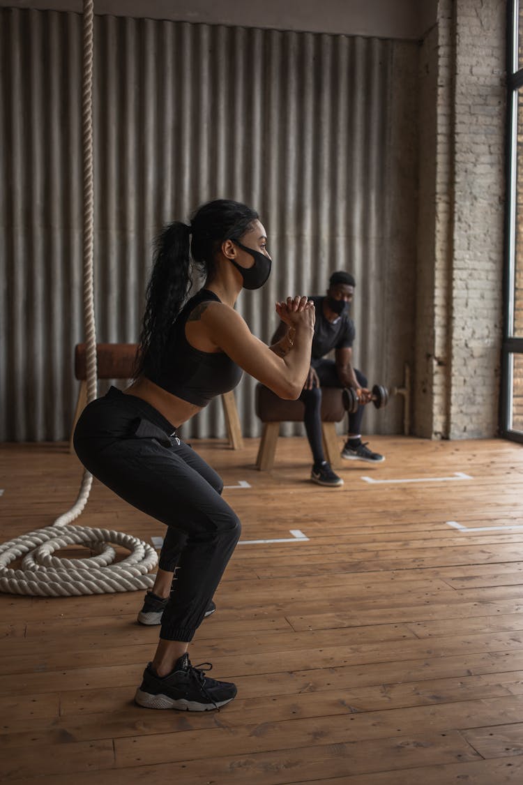 Black Sportswoman Squatting Against Partner With Dumbbell In Gymnasium