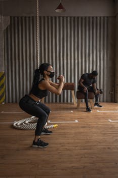 Black sportswoman in fabric mask squatting on floor against anonymous male athlete with dumbbell during training in gymnasium