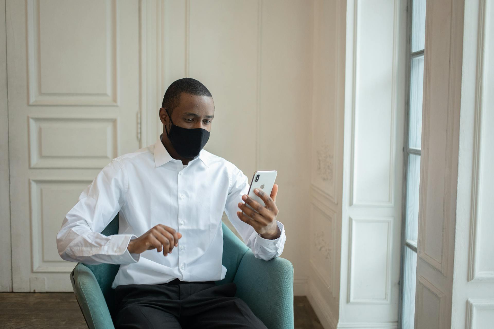 Black male executive in white shirt and fabric mask sitting in armchair while talking on cellphone during video chat indoors