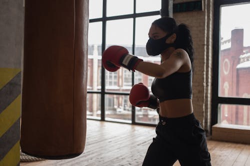Determined African American female fighter in cloth face mask and gloves during workout against boxing bag in gym