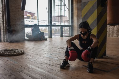 Free Exhausted African American female fighter in boxing gloves and cloth face mask sitting on parquet against unrecognizable partner in gymnasium Stock Photo