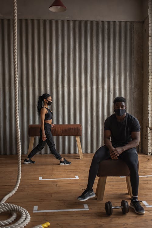 Ethnic male athlete sitting on pommel horse against sportswoman in cloth face mask working out in gymnasium