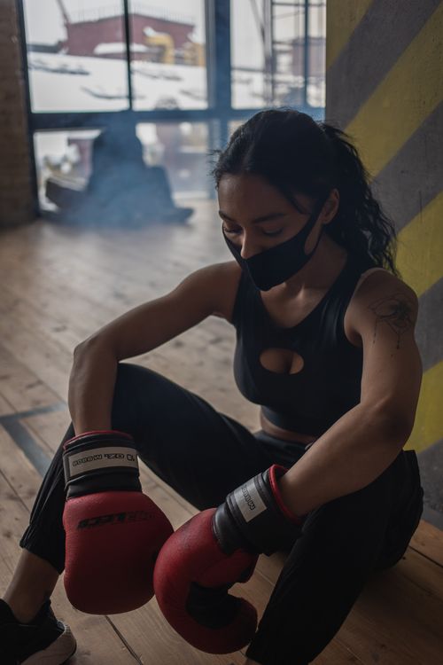 Free Tired African American female athlete in boxing gloves and fabric mask resting on floor against anonymous partner in gymnasium Stock Photo