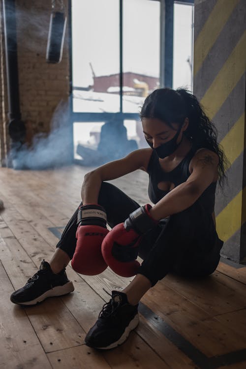 Free Exhausted black sportswoman in boxing gloves and cloth face mask having break from training on floor in gymnasium Stock Photo