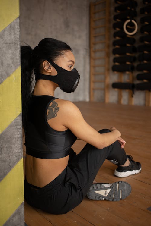 Back view of African American female athlete in sportswear and protective respirator sitting on floor in fitness studio