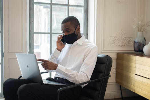 Businessman in mask talking on phone while using laptop indoors. Modern remote work setup.