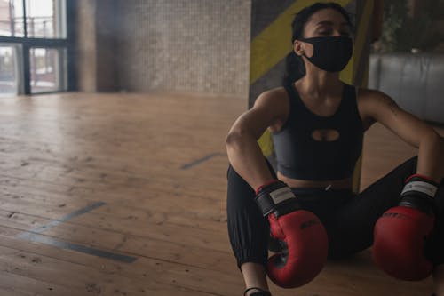 African American female boxer in protective mask resting on floor after training in fitness center