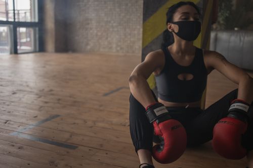 Free African American female boxer in protective mask resting on floor after training in fitness center Stock Photo