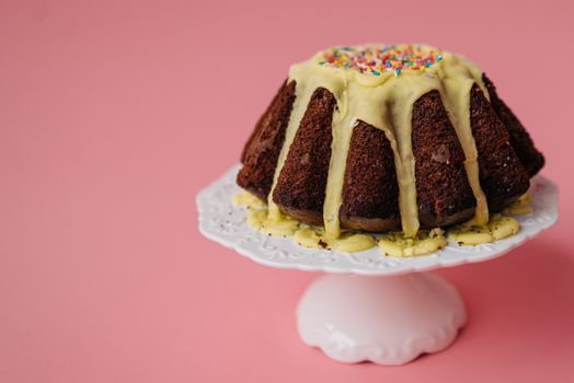 A chocolate bundt cake with creamy icing and sprinkles on a stand against a pink background.