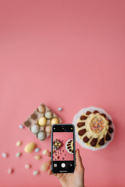 Person Taking Picture of Easter Eggs and Cake on Pink Background
