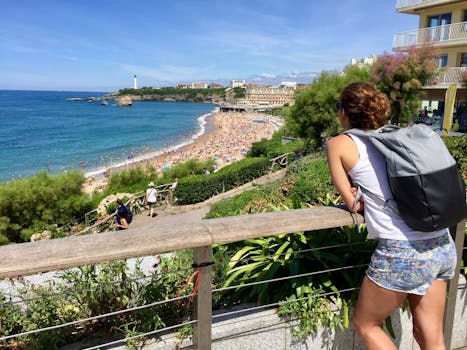 A young woman with a backpack gazes over the bustling beach of Biarritz, France on a sunny day. by Monica Silvestre