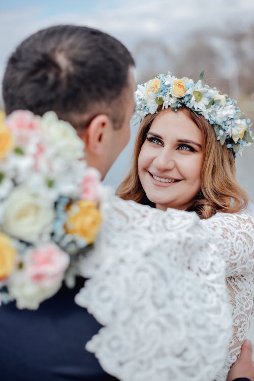 Loving bride in white dress and wreath smiling happily while embracing groom at wedding ceremony