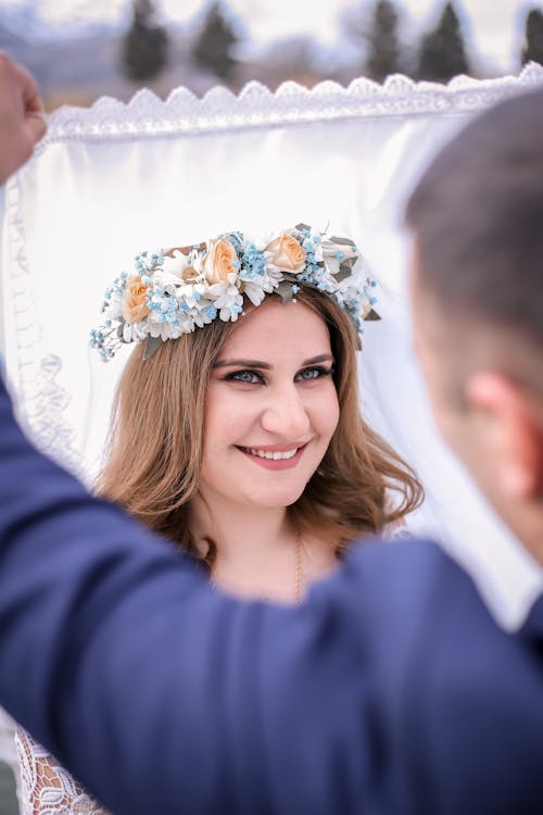 Crop groom lifting veil of positive bride in floral wreath during romantic wedding ceremony in nature