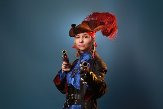 Young self assured woman in hat with feathers and vintage pistols looking at camera on gray background