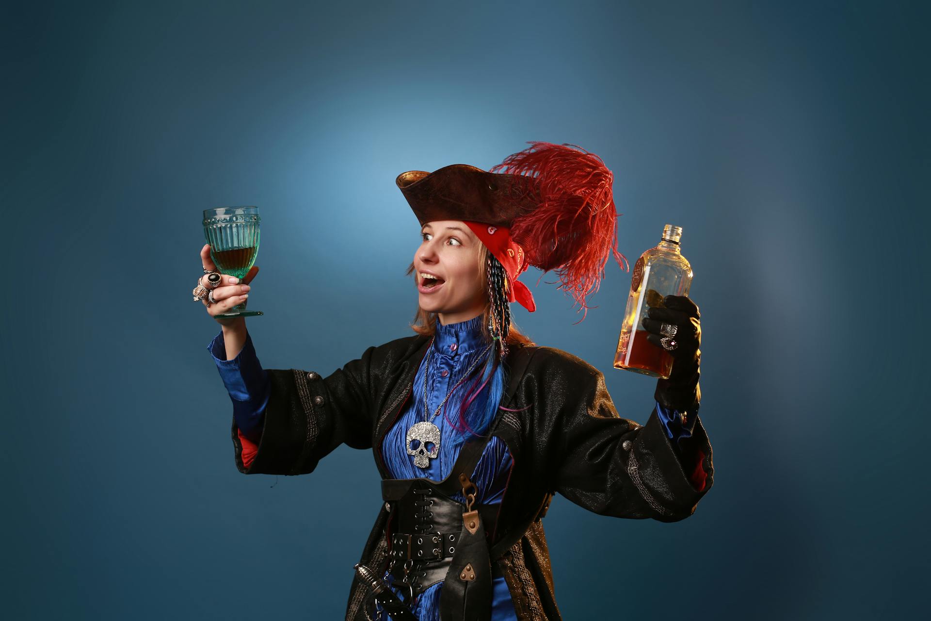 Amazed woman in pirate costume and pendant with decorative skull holding vintage glass and bottle of booze