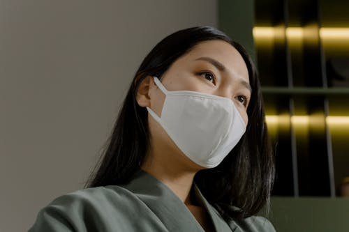 Young Asian female with long dark hair in medical mask and gray jacket looking away against white wall