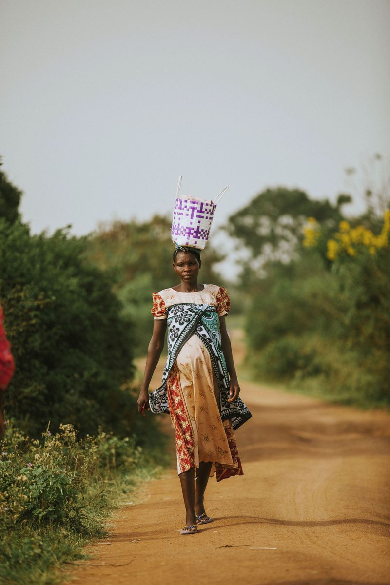 A pregnant African woman carrying a basket on her head while walking along a rural dirt road.