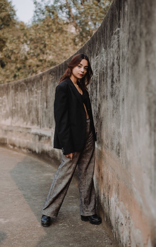 Free Full body of young female model wearing gray trousers and black jacket standing at concrete fence Stock Photo