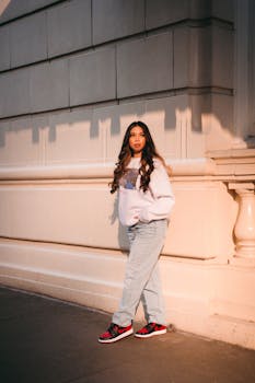 Young woman in jeans and sweatshirt posing against an elegant building at sunset.