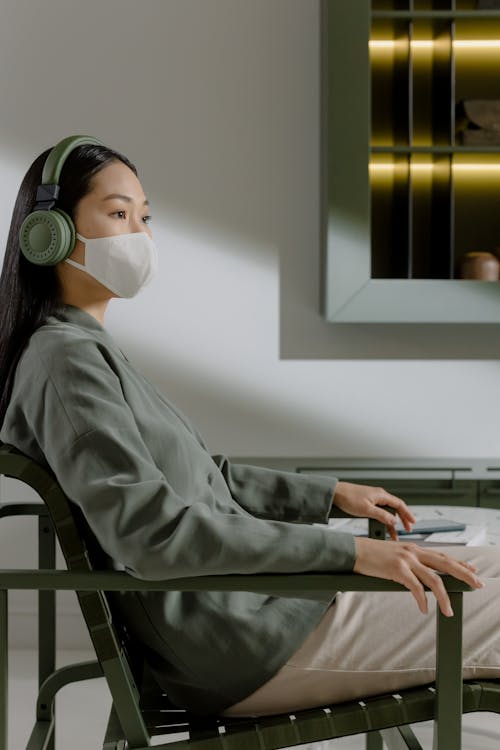 A Woman with Face Mask Sitting on a Chair while Listening to Music on a Wireless Headphones