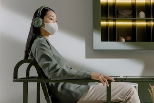 A Woman with Face Mask Sitting on a Chair while Listening Music on a Wireless Headphones