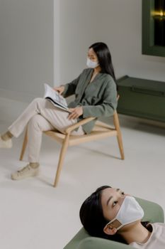 Two women wearing masks and relaxing indoors, maintaining social distance.