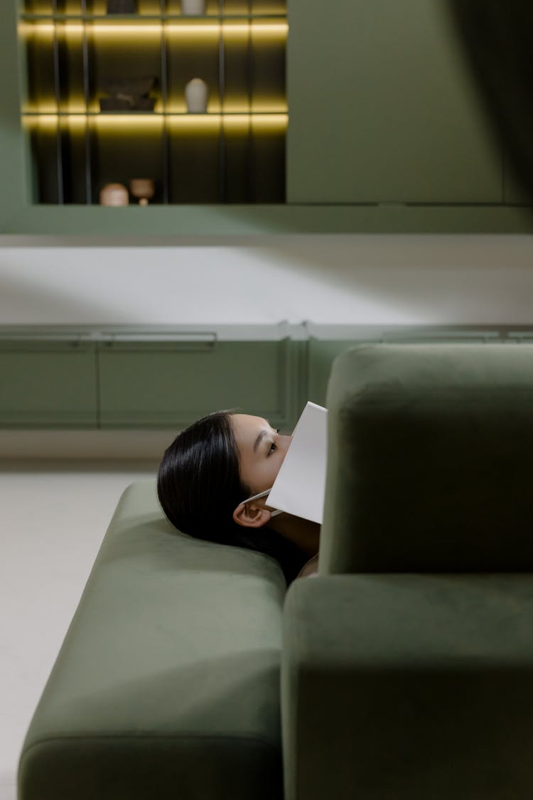 Woman Laying Down On Couch Covering Mouth With A Book